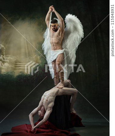 Divine Judgment. Man with angel wings stands over another man, holding sword aloft as light beams shine through clouds against dramatic background. 115848931
