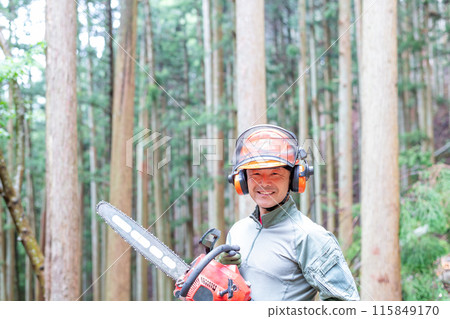 A forest worker with a chainsaw and a smile facing forward with a well-maintained forest in the background 115849170
