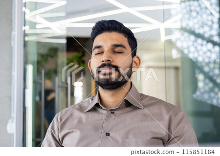 A calm man with his eyes closed is meditating in a contemporary office environment. This serene moment captures the essence of stress relief and mindfulness practice in a work setting 115851184