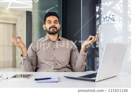 A businessman sitting at his desk in an office setting, practicing meditation with a calm expression. He aims to find relaxation and reduce stress while working. 115851186
