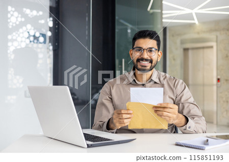 A smiling man in an office setting, enthusiastically opening and reading a letter at his desk, with a laptop and notebook in view. The scene conveys positivity, achievement, and professional success. 115851193