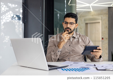 A focused financier or accountant uses a calculator and tablet while analyzing data on a laptop in a contemporary office. The person appears deep in thought, emphasizing finance, business analysis 115851205