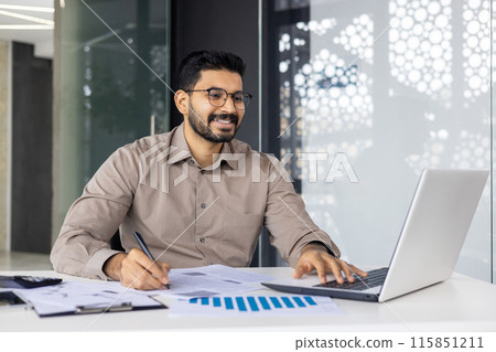 Confident businessman smiling while working on laptop and analyzing documents at a modern office. Concept of productivity, professionalism, and business success in a contemporary workspace. 115851211