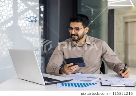A confident financier uses a calculator and a laptop in a modern office setting. The image depicts a professional handling financial tasks with focus and concentration. 115851216
