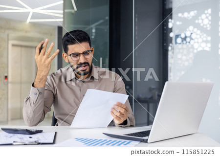 Young man wearing glasses and looking stressed and frustrated while holding paperwork in an office setting. He is seated at a desk with a laptop, documents. Emotion of worry and confusion. 115851225