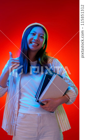 Cool study. Successful education. Satisfied cheerful girl with books showing thumb up like gesture in blue color neon light isolated on red copy space background. Cool study. Successful education. Satisfied cheerful girl with books showing thumb up like gesture in blue color neon light isolated on red copy space background. 115851312