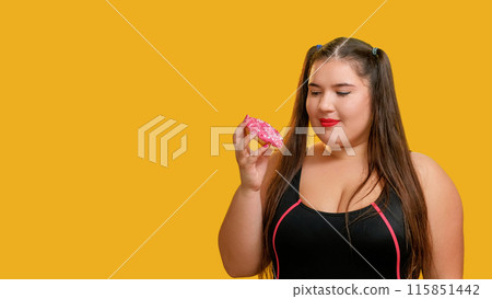 Unhealthy food. Sweet dessert. Strawberry doughnut. Hungry chubby overweight woman looking at desirable pink donut with anticipation delicious isolated on yellow empty space colorful background. Unhealthy food. Sweet dessert. Strawberry doughnut. Hungry chubby overweight woman looking at desirable pink donut with anticipation delicious isolated on yellow empty space colorful background. 115851442