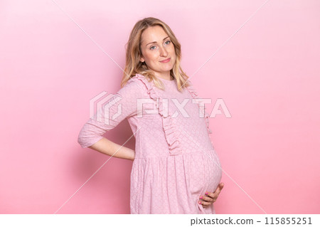 Pregnant woman in a pink dress holding her belly against a pink background 115855251