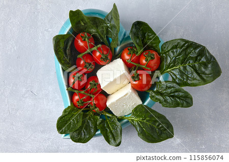 Cherry tomatoes, brine cheese and spinach in a blue decorative bowl on a gray stone table. Top view with a copy of the space 115856074