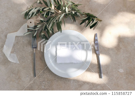 Summer wedding stationery. Blank business card, invitation on white ceramic plate. Olive tree branches, silver fork, knife. Festive table setting. Beige marble background in sunlight. Flat lay, top. 115858727