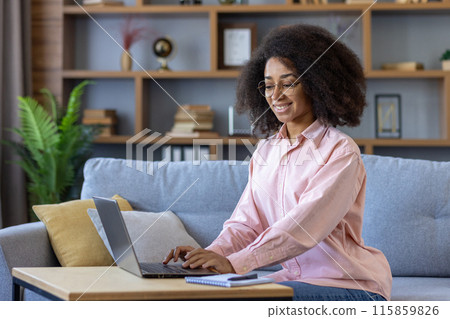 Smiling woman working from home on laptop seated on sofa in cozy living room. Concept of remote work, home office, comfort Smiling woman working from home on laptop seated on sofa in cozy living room. Concept of remote work, home office, comfort 115859826