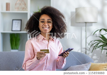 Smiling woman holding credit card and smartphone indoors. Concept of online shopping, digital payment, and e-commerce at home in a cozy living room setting 115859860