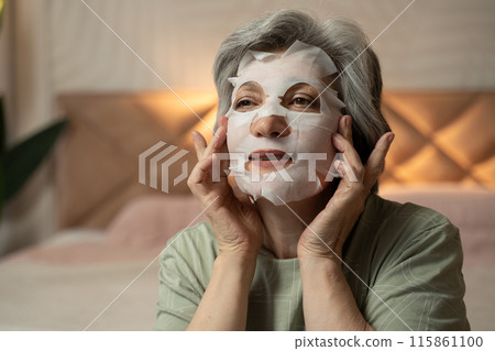 An elderly woman puts a cosmetic mask on her face in the evening in her bedroom. The soft and warm lighting creates a relaxing atmosphere. 115861100