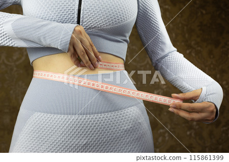 Girl in workout suit with cutout for her belly in gym measures her waist with special flexible tape, concept of sports and healthy lifestyle Girl in workout suit with cutout for her belly in gym measures her waist with special flexible tape, concept of sports and healthy lifestyle 115861399