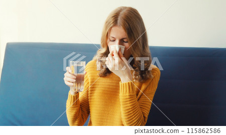 Sick exhausted woman sneezing blow nose using tissue drinks water from a glass taking a medication 115862586