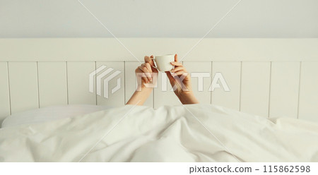 Cheerful lazy woman waking up after sleeping lying in bed holding cup in her hands asks for coffee 115862598