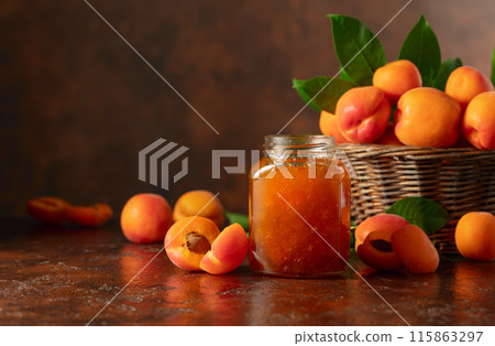 Apricot jam in glass jar and fresh fruits on a brown table. 115863297