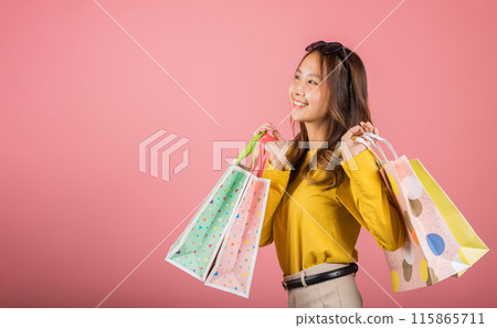 A stylish Asian lady, elegantly holding shopping bags, smiles with joy in a studio portrait on a pink background with copy space. She's the epitome of happy shopping. black friday 115865711