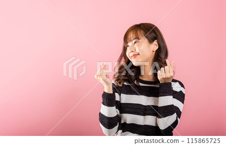 A confident woman stands with a smile, creating a mini heart sign with her finger on a pink background. Asian portrait of a beautiful young female sending love for Valentine's Day in a studio shot. A confident woman stands with a smile, creating a mini heart sign with her finger on a pink background. Asian portrait of a beautiful young female sending love for Valentine's Day in a studio shot. 115865725
