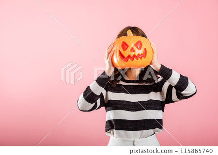 A portrait of a joyful Asian woman holding model pumpkins on her head in a Halloween-themed studio shot. She's all smiles with ghost pumpkins on a pink background. A portrait of a joyful Asian woman holding model pumpkins on her head in a Halloween-themed studio shot. She's all smiles with ghost pumpkins on a pink background. 115865740