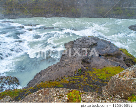 Tourists standing on the rocky edge observing the powerful flows of Gullfoss, one of Icelands iconic waterfalls, against a backdrop of rugged landscape. Iceland Tourists standing on the rocky edge observing the powerful flows of Gullfoss, one of Icelands iconic waterfalls, against a backdrop of rugged landscape. Iceland 115866892