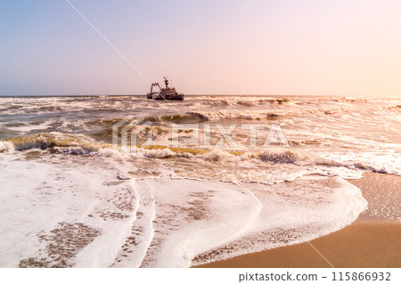A shipwreck is embraced by the turbulent waters of the Skeleton Coast, under the warm hues of a fading sunset. 115866932
