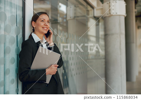 Happy businesswoman in a suit by an office window, talking on the phone and holding a tablet 115868034