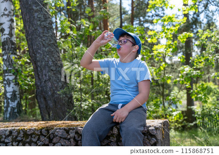 A boy on stone wall in forest drinks water, sun through trees 115868751