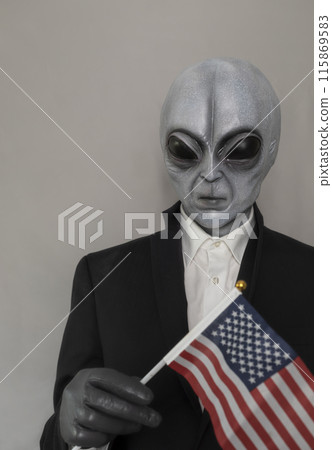 Extra Terrestrial Being, Portrait Of Alien Wearing Suit And Holding American Usa Flag on Gray Background. World UFO Day, Unidentified Flying Objects. Vertical Plane. Grey Humanoid. 115869583