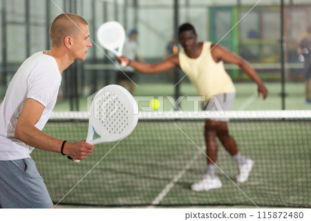 Padel player playing padel in a padel court indoor behind net Padel player playing padel in a padel court indoor behind net 115872480