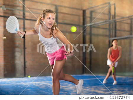 Expressive energetic girl playing padel on indoor court 115872774