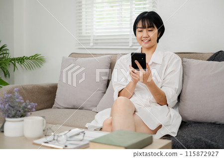 Young woman sitting on a sofa using a smartphone in a bright, modern living room with minimalist decor and natural light. Young woman sitting on a sofa using a smartphone in a bright, modern living room with minimalist decor and natural light. 115872927