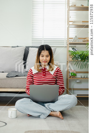 Young woman sitting cross-legged on the floor, working on a laptop in a cozy, modern living room with natural light. Young woman sitting cross-legged on the floor, working on a laptop in a cozy, modern living room with natural light. 115872938