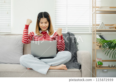 Young woman in casual clothes celebrating success while working on a laptop at home, sitting on a sofa in a bright and modern living room. Young woman in casual clothes celebrating success while working on a laptop at home, sitting on a sofa in a bright and modern living room. 115872939