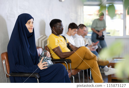 Young asian woman in burqa together with other people of different nationality waiting in office Young asian woman in burqa together with other people of different nationality waiting in office 115873127