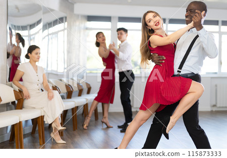 Smiling young european woman in red clothes dancing latin dance with African american man in elegant clothes, energetic and fast tango in dance salon Smiling young european woman in red clothes dancing latin dance with African american man in elegant clothes, energetic and fast tango in dance salon 115873333