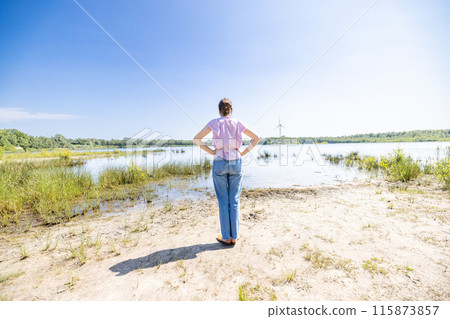 A woman stands by the lakeside, gazing at a serene landscape of a wind turbine in the background 115873857