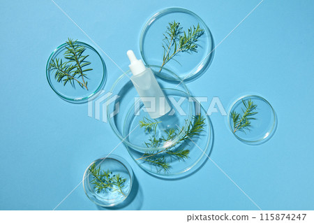 Blue background features some petri dishes containing tea tree leaves and an unlabeled bottle of serum. Empty label for cosmetic product mockup 115874247