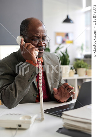 Vertical portrait of Black senior man using landline phone and talking business at workplace in office Vertical portrait of Black senior man using landline phone and talking business at workplace in office 115878372