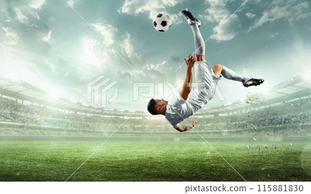 Panoramic view of performs impressive kick, caught mid-air with ball high above him against stadium illuminated by spotlights. 115881830
