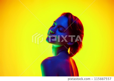 Beautiful young woman with short brown hair looks over shoulder in neon light against gradient studio background. 115887557