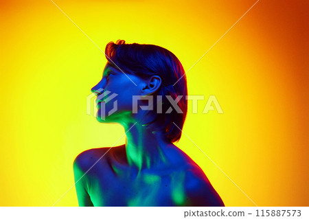 Young woman with short brown hair is bathed in neon light highlighting her skin's health and radiance against gradient studio background. 115887573
