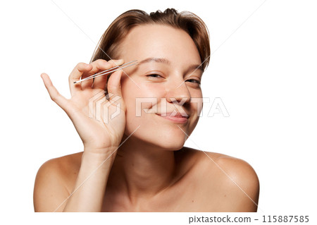 Young woman with short brown hair smiles while holding tweezers near her eyebrow, as if grooming it against white studio background. 115887585