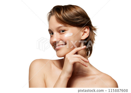 Woman with short brown hair smiles applying lotion to her cheek against white studio background. Morning routine. 115887608
