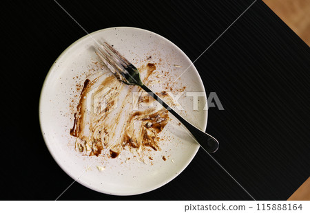 Top view empty fork and dish with stains of chocolate and cake after eating. Food dish is on black table with space. Top view empty fork and dish with stains of chocolate and cake after eating. Food dish is on black table with space. 115888164