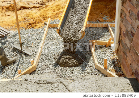 Construction worker pours cement on side of house to create new sidewalk that will stretch along side building 115888409