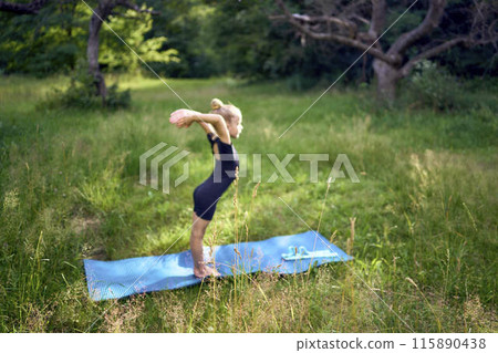 little 5-year-old girl in a black minimalist overall does gymnastics in the garden, outdoors 115890438