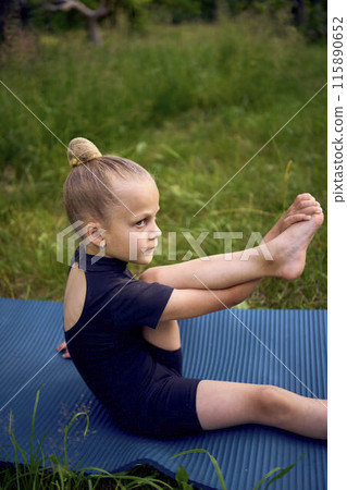 little 5-year-old girl in a black minimalist overall does gymnastics in the garden, outdoors 115890652