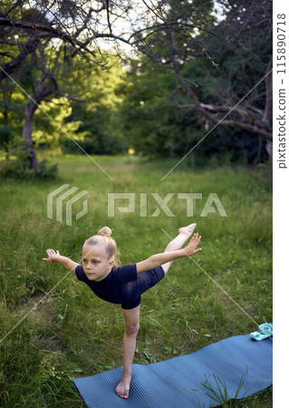 a little 5-year-old girl in a black minimalist jumpsuit does gymnastics in the garden, outdoors 115890718