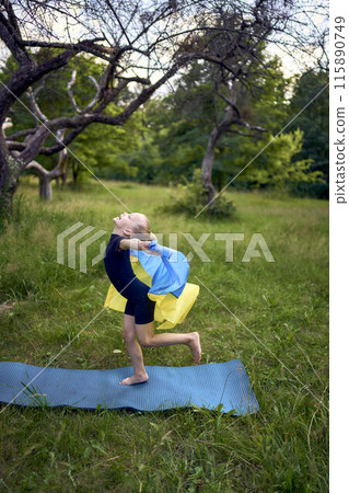a little 5-year-old gymnast in a black minimalist jumpsuit is practicing with the flag of Ukraine in the garden 115890749